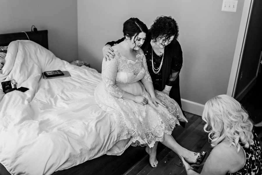 A bride puts on her wedding shoes with the help of her mother and bridesmaid.